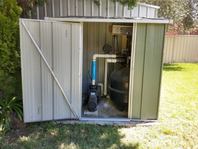 Metal pool equipment shed in the backyard hiding the pool equipment. The pool equipment enclosure contains a pool pump and sand filter mounted on concrete pad.
